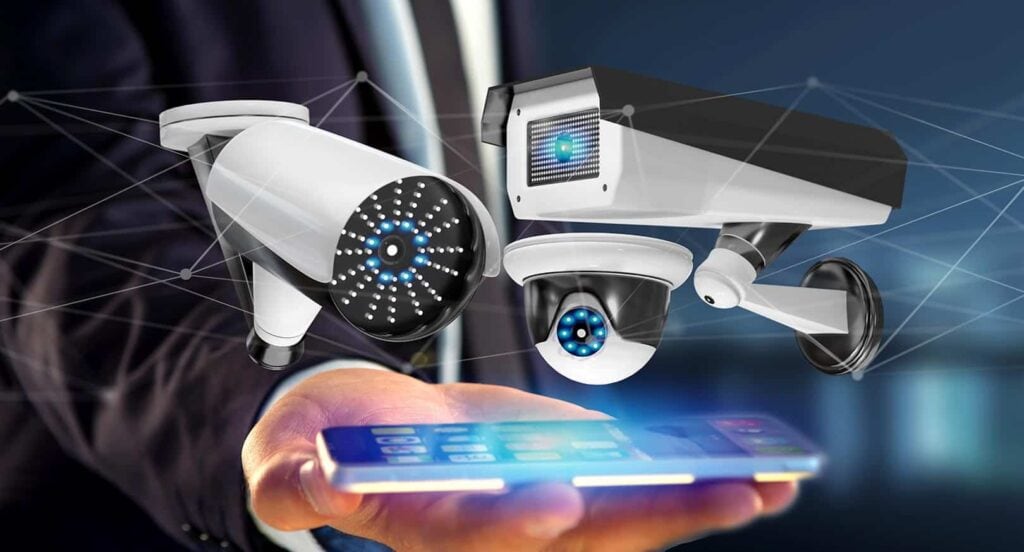 A person in a suit holds a smartphone with holographic images of three security cameras above it, symbolizing how smart surveillance technology benefits a general contractor in Houston, TX.