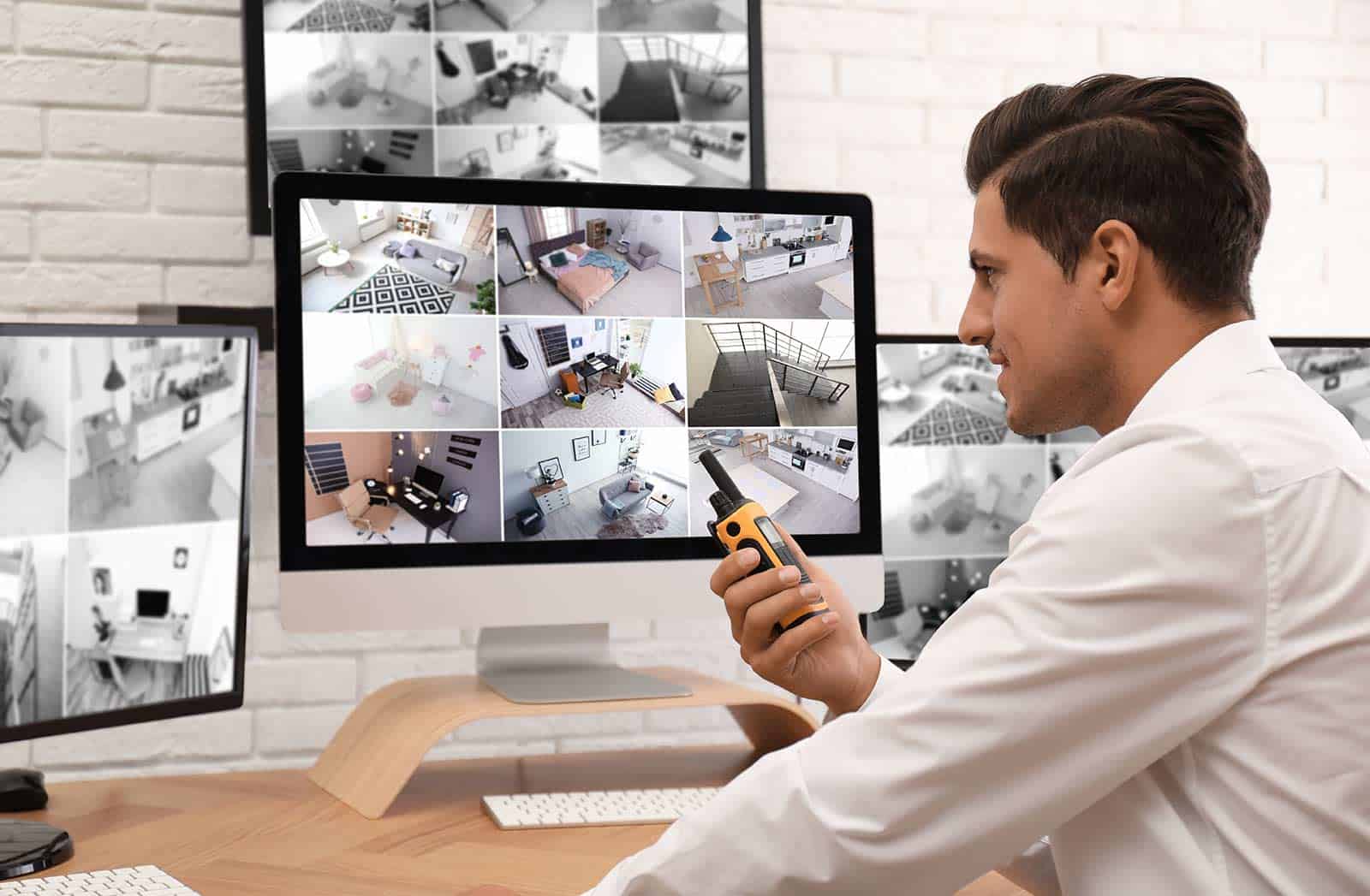 A man in a white shirt monitors multiple security camera feeds on several computer screens while holding a walkie-talkie in an office setting for a general contractor Houston, TX.