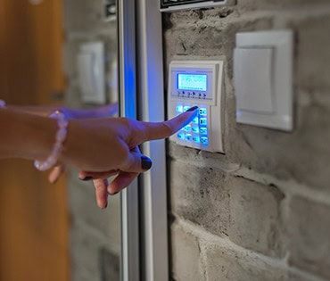 A person’s hand is entering a code on a wall-mounted electronic security keypad with blue backlighting, installed by a general contractor in Houston, TX, and attached to a brick wall next to a light switch.