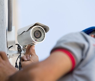 A general contractor in Houston, TX, wearing a gray shirt, is installing or adjusting a security camera on a white wall, ensuring the lens faces outward and the cables are properly connected.