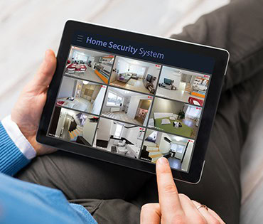 A person holds a tablet displaying a home security system interface with multiple live camera feeds, showcasing different rooms and areas—a smart solution recommended by a general contractor in Houston, TX.