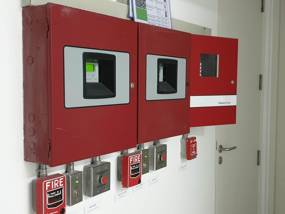 Three red fire alarm control panels and manual pull stations are mounted on a white wall near a closed door; one panel’s door is open, revealing its controls, installed by a general contractor in Houston, TX.