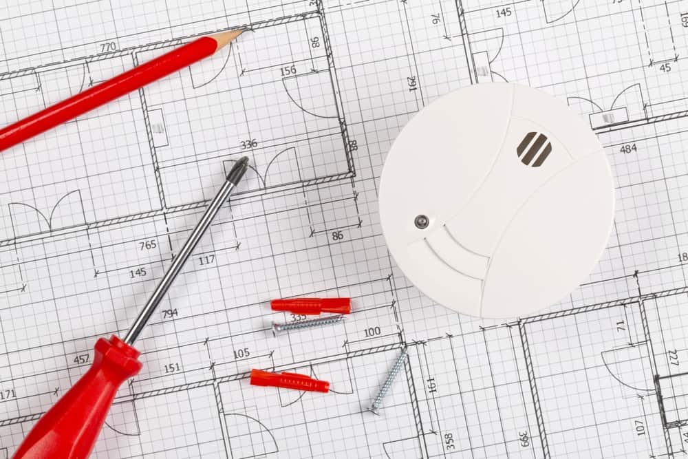 A red pencil, red screwdriver, screws, wall plugs, and a white smoke detector are arranged on a technical floor plan drawing—a scene familiar to any general contractor Houston trusts for precision and expertise.