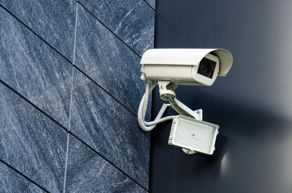 A white security camera and a rectangular floodlight are mounted on the exterior wall of a building with dark, diagonally tiled panels—installed by a trusted general contractor in Houston, TX.