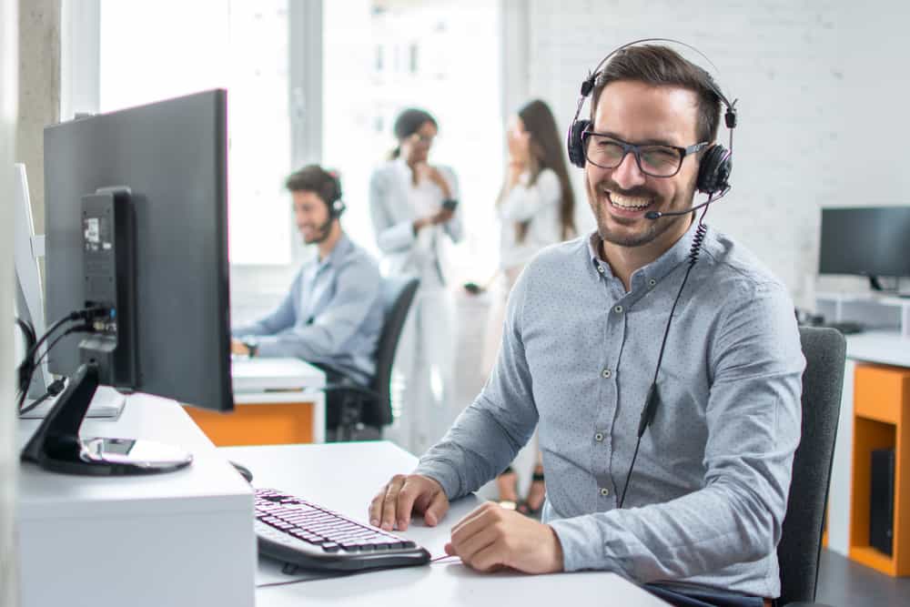A smiling man with glasses and a headset sits at a desk with a computer in a modern office, representing a general contractor Houston, TX, while other people work and talk in the background.