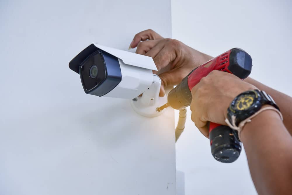 A person uses a red electric drill to install a security camera on a white wall, securing the camera's mounting bracket with screws—work often handled by a general contractor Houston, TX trusts for reliable installations.