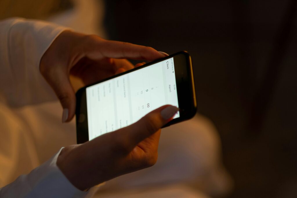 A person holds a smartphone in both hands, interacting with a bright screen displaying text and numbers, possibly using an app related to commercial security systems Houston. The background is dimly lit and out of focus.