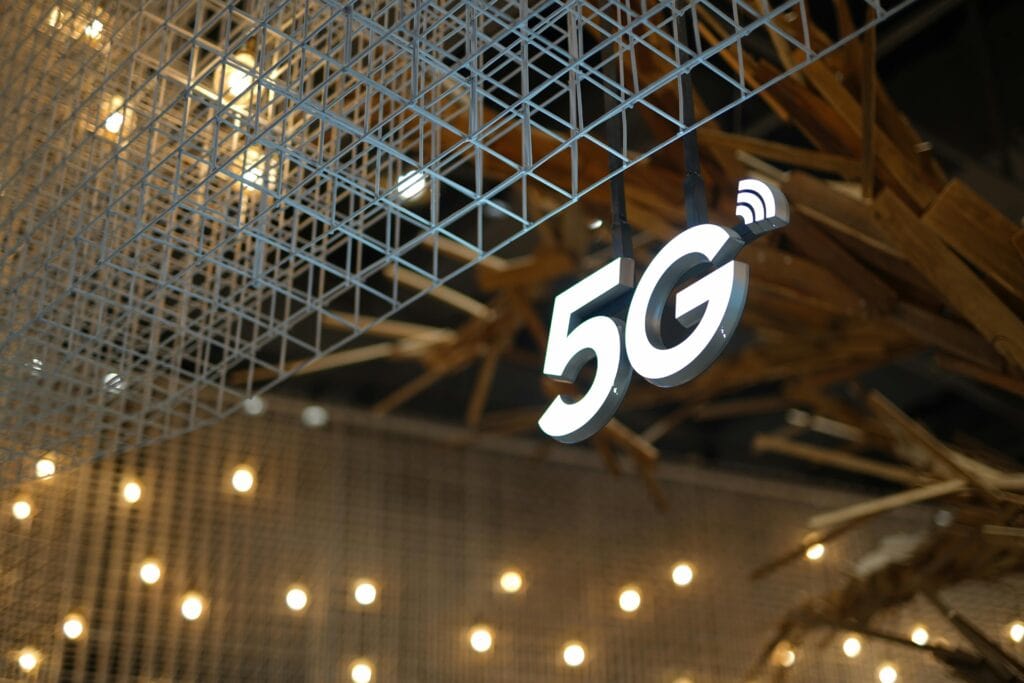 Illuminated "5G" sign with a wireless signal icon hangs from a grid-like metal structure, evoking cutting-edge tech like residential security systems Houston, with blurred lights and abstract wooden elements in the background.